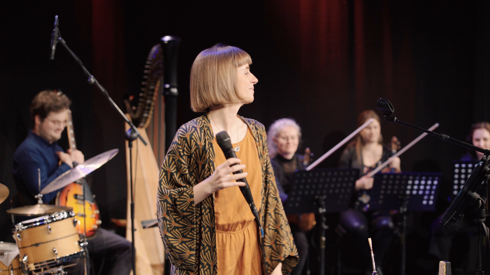 Miriam Hanika auf der Bühne des Saal X, Gasteig HP8 mit ihrem großen Ensemble im Hintergrund.